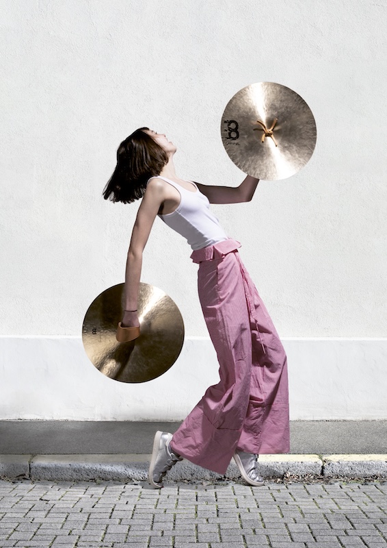 Femme en mouvement tenant deux cymbales, l’une devant elle et l’autre derrière, sur fond de mur blanc, dans une posture dynamique évoquant la musique et le rythme